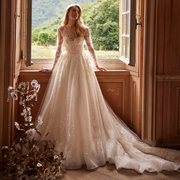 Shining long sleeve wedding dress with a sheer illusion neckline, beaded lace bodice, and a voluminous glitter tulle A-line skirt with a cascading train by a window.