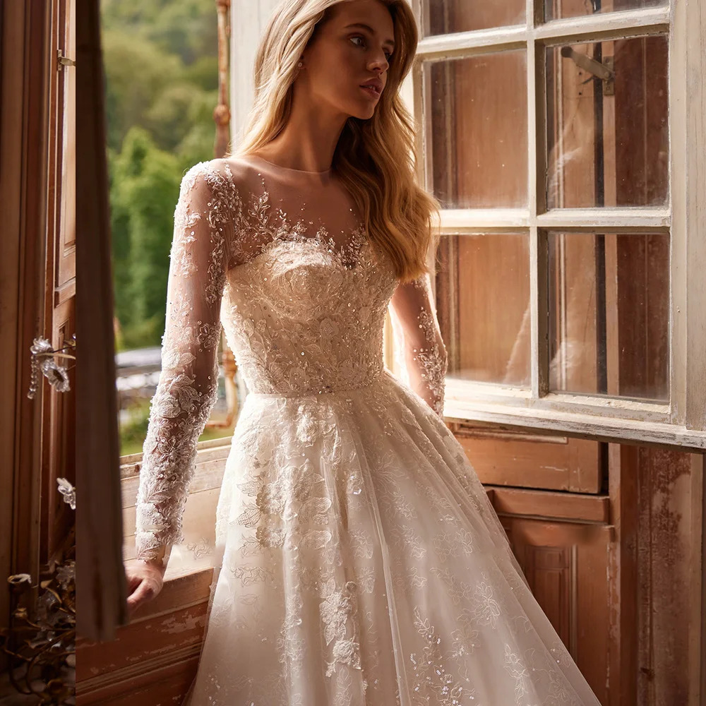 Elegant bride posing by a rustic window in a sparkly long sleeve A-line wedding gown with sheer embroidered floral sleeves.