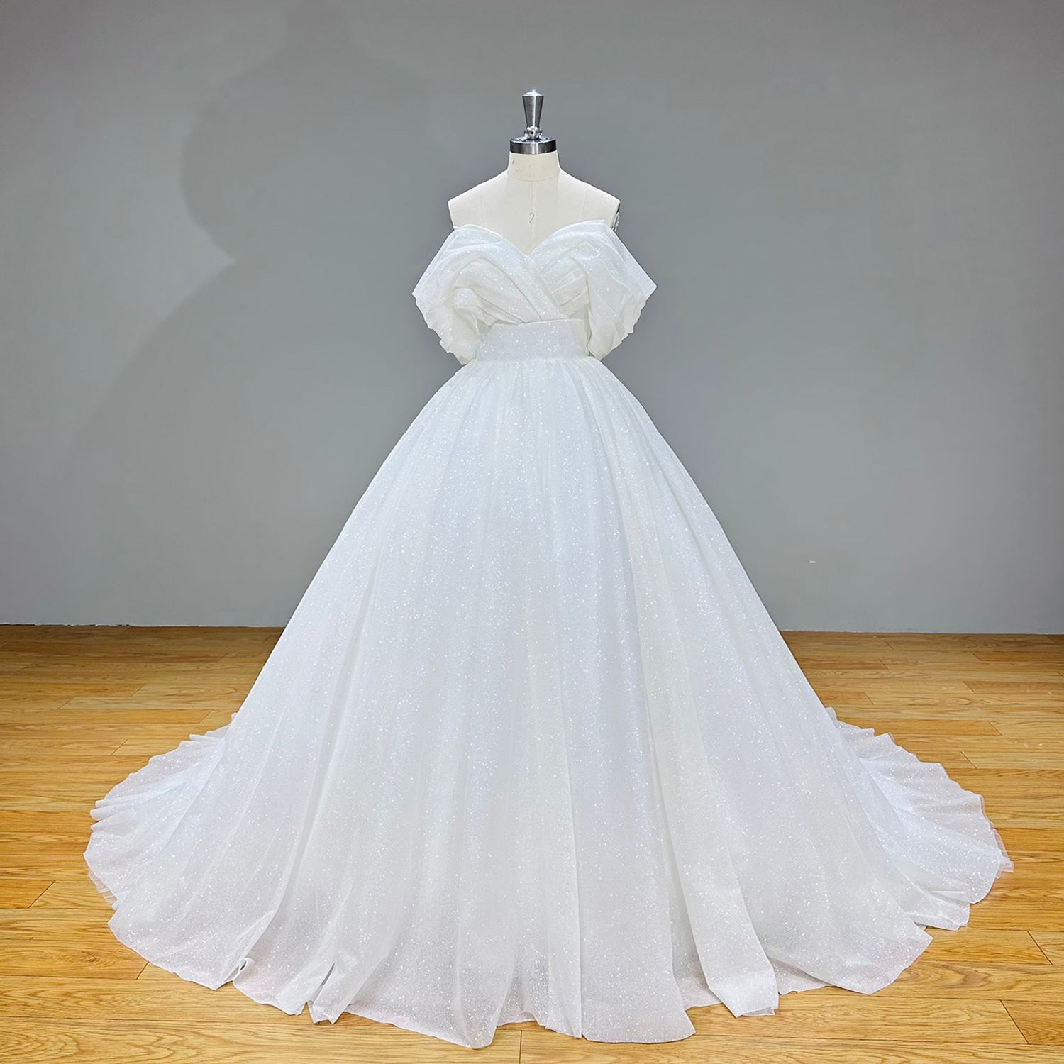 Full front product photography of the shining off-the-shoulder ball gown wedding dress with a voluminous pleated skirt against a gray studio backdrop.
