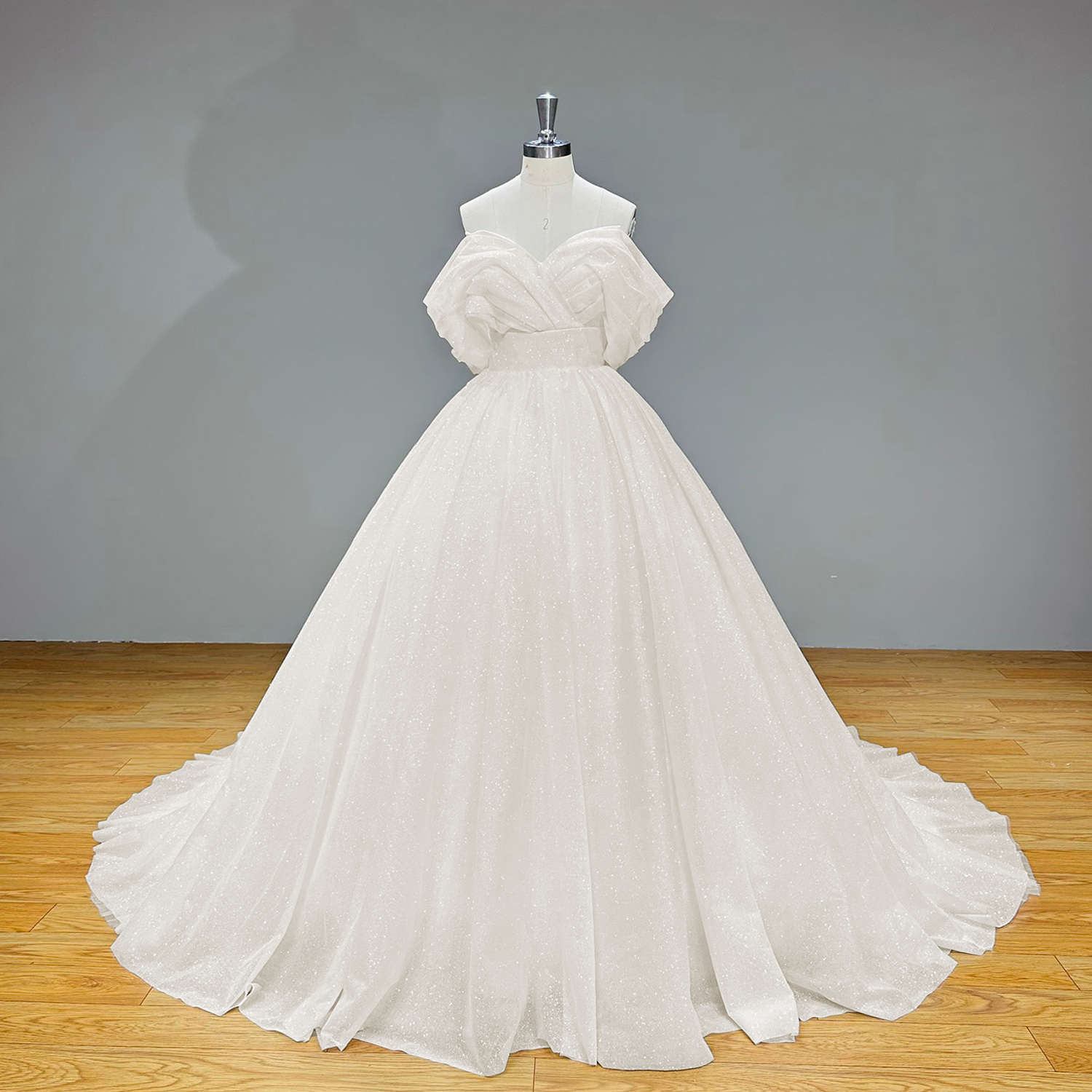 Full front product photography of the shining off-the-shoulder ball gown wedding dress with a voluminous pleated skirt against a gray studio backdrop.