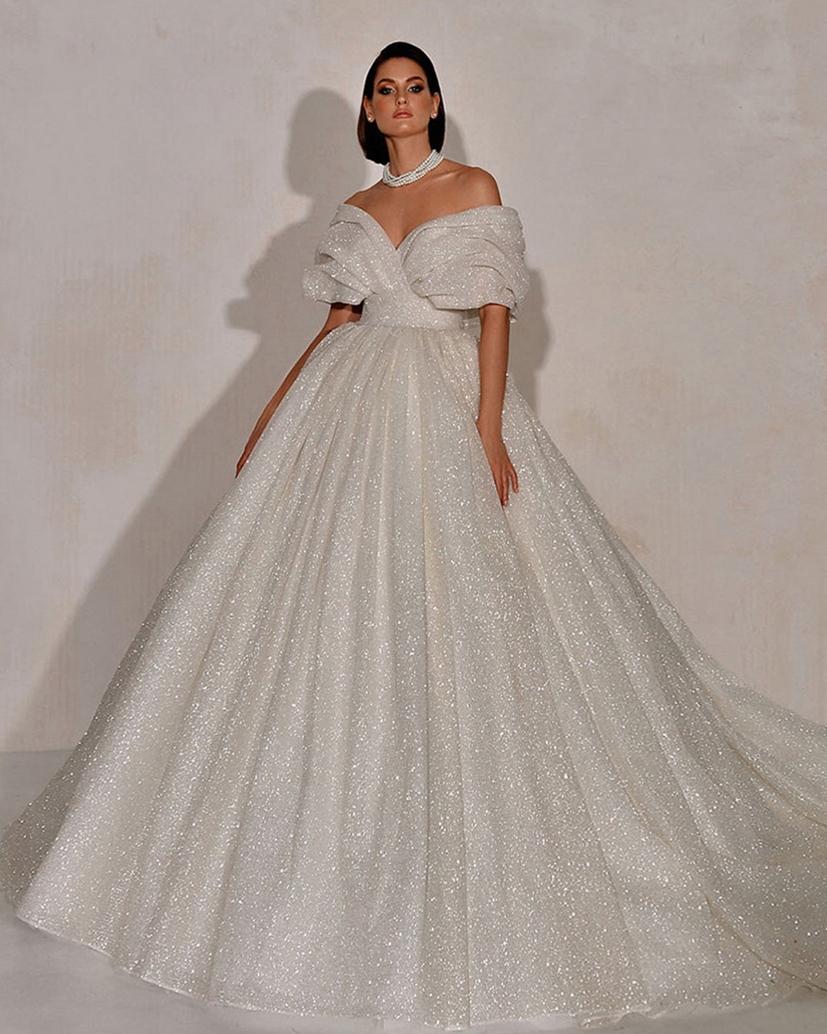 High-fashion bridal look featuring a model in a glittering off-the-shoulder ball gown wedding dress with structured pleated sleeve drapes and a pearl choker.