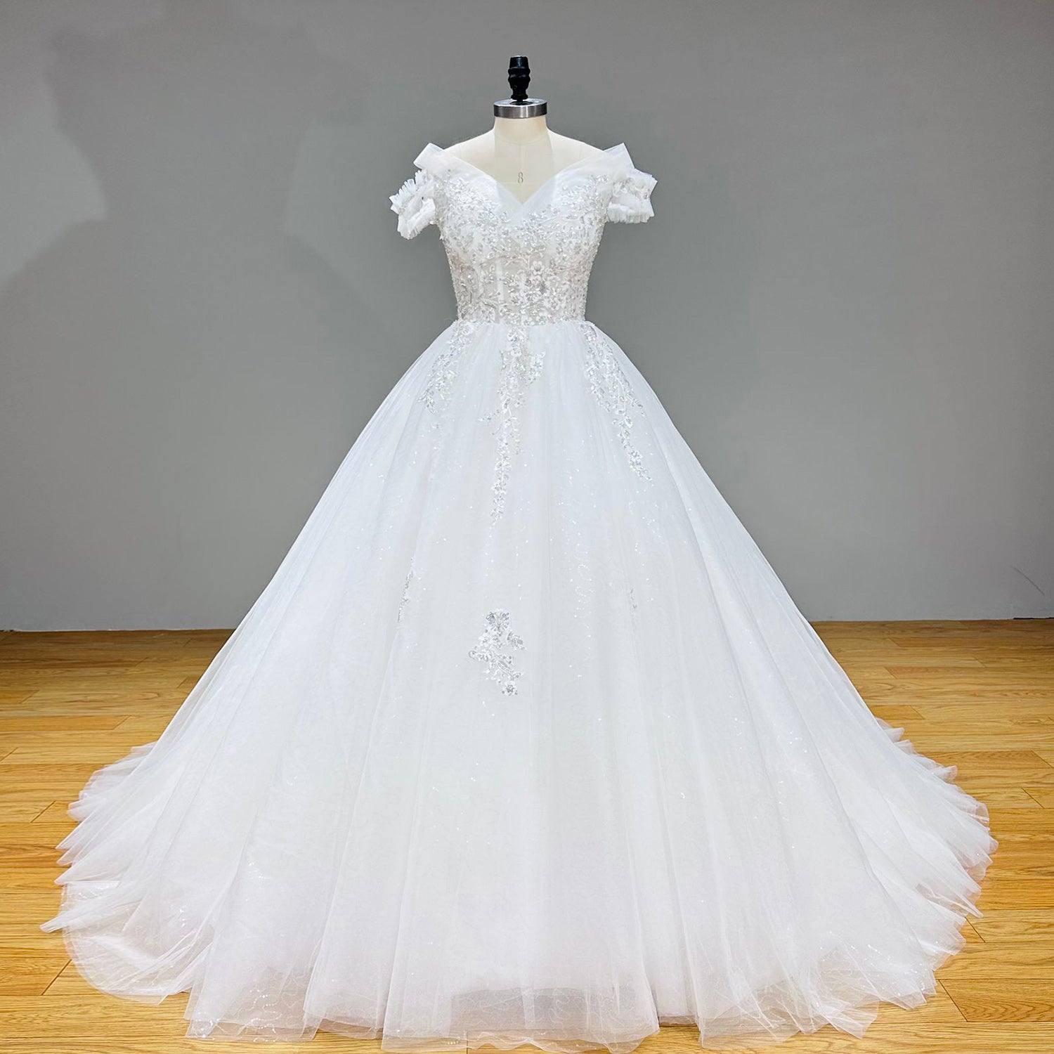 Full-length studio front view of a majestic white glitter tulle princess ball gown wedding dress with ruffled off-the-shoulder straps on a dress form.
