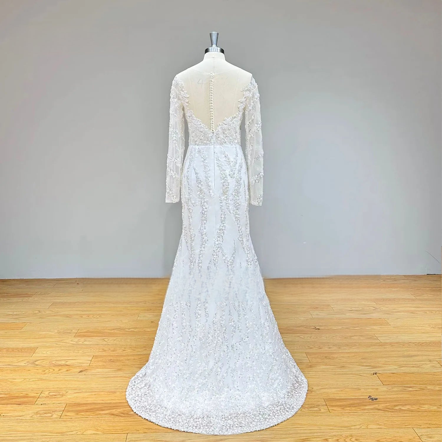 Rear perspective of the long sleeve mermaid wedding dress on a mannequin highlighting the sheer back with pearl buttons and detailed sequin patterns.