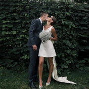 Modern short satin V-neck wedding dress featuring a sleeveless mini sheath silhouette and a dramatic detachable trailing back bow, styled for an outdoor elopement.