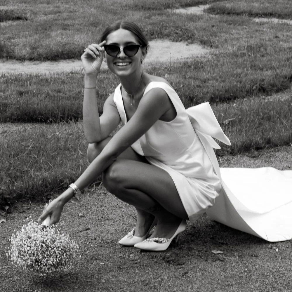 Elegant short white satin bridal gown with a low back and removable train, styled playfully with sunglasses and embellished kitten heels for a casual reception.