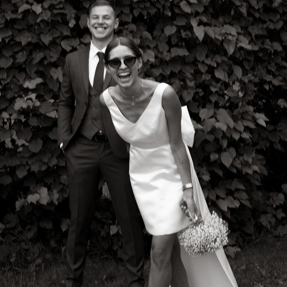 Minimalist sleeveless white satin bridal mini dress featuring a tailored crossover V-neckline, styled with a simple baby's breath wedding bouquet.