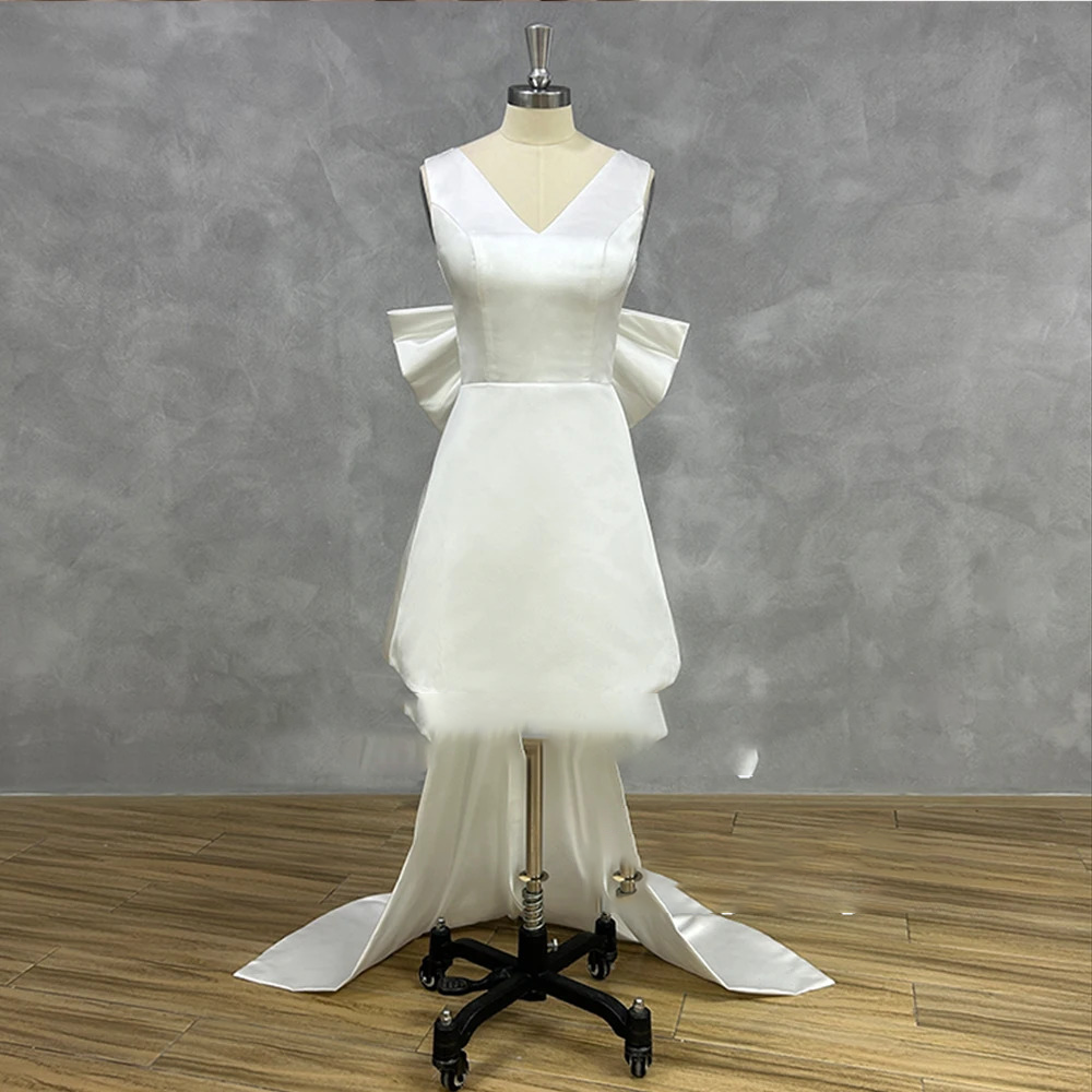 Short satin V-neck wedding dress on a mannequin featuring a sleek sleeveless mini sheath bodice, a tailored waistline, and a dramatic detachable cascading back bow train.