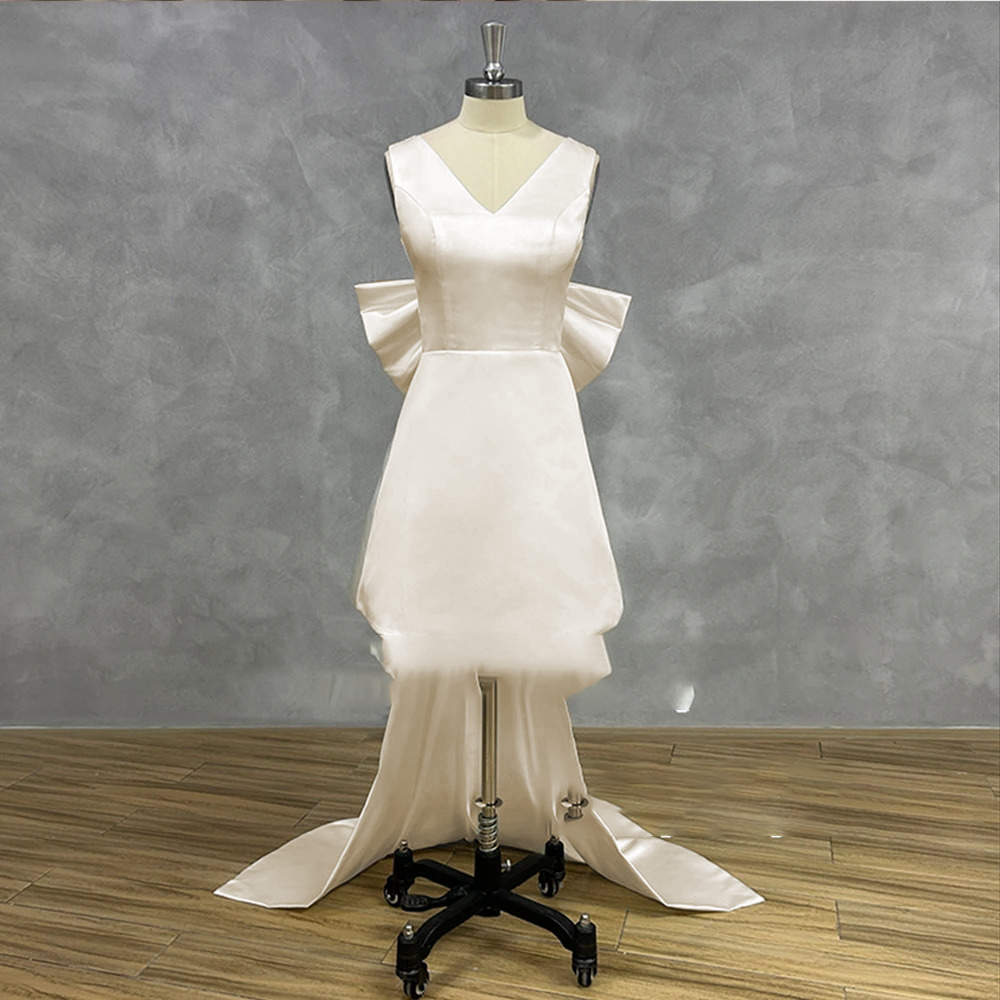 Soft ivory short satin bridal gown on a mannequin showcasing a classic V-neckline, structured princess seams, and a removable floor-length statement bow overskirt train.