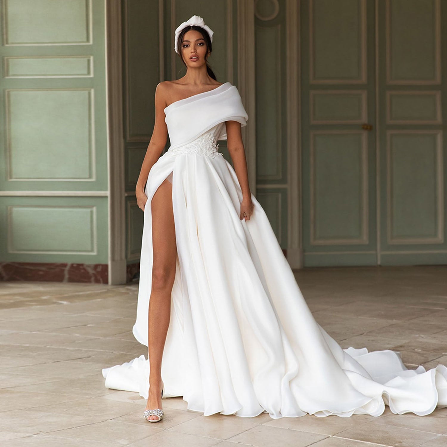 Glamorous bride wearing a modern one shoulder organza wedding dress with a dramatic thigh-high slit skirt and an embroidered floral waistband, posing in an elegant chateau hall.