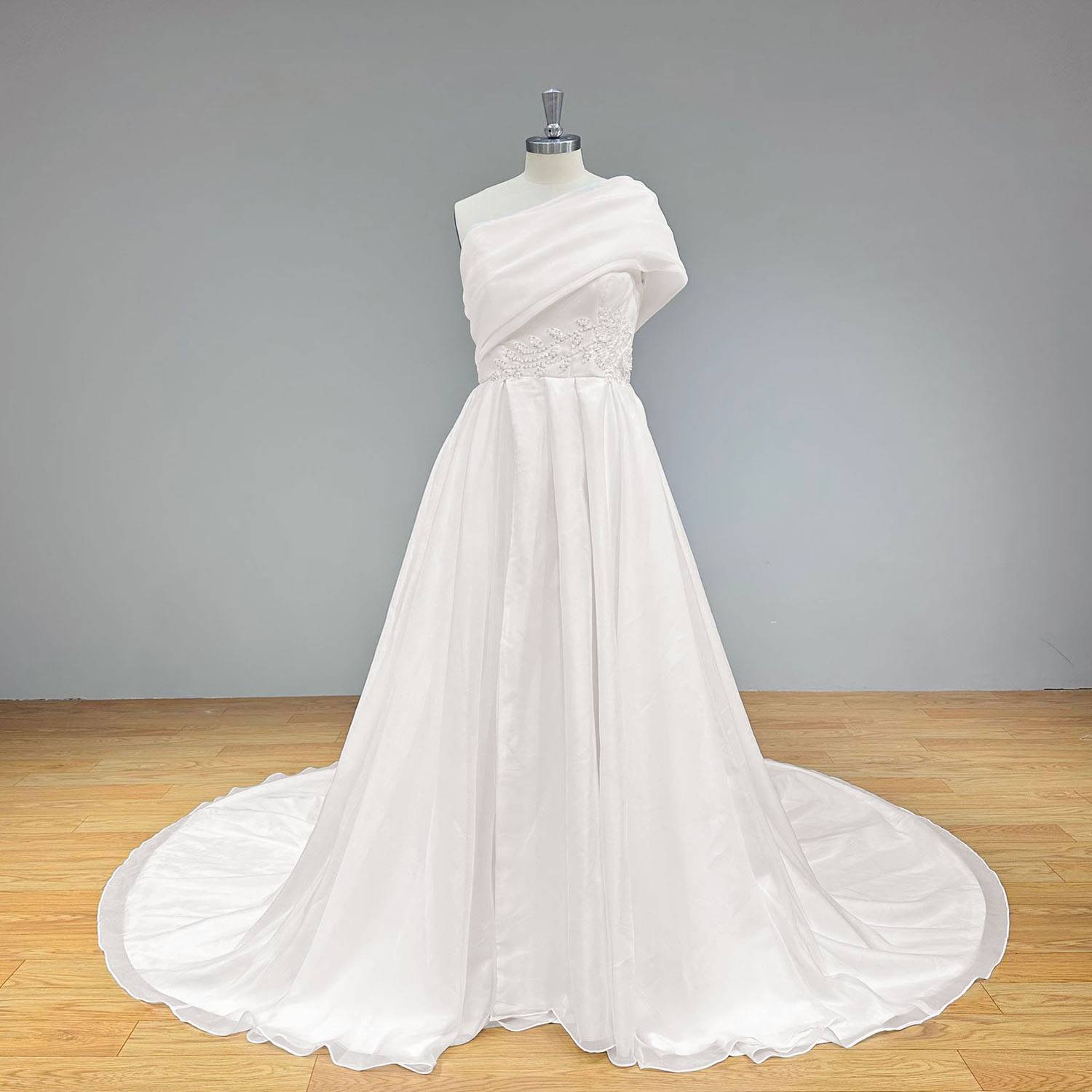 Studio front shot of the high-slit organza wedding dress on a dress form against a solid gray backdrop, highlighting the light-reflective layers of the romantic skirt.