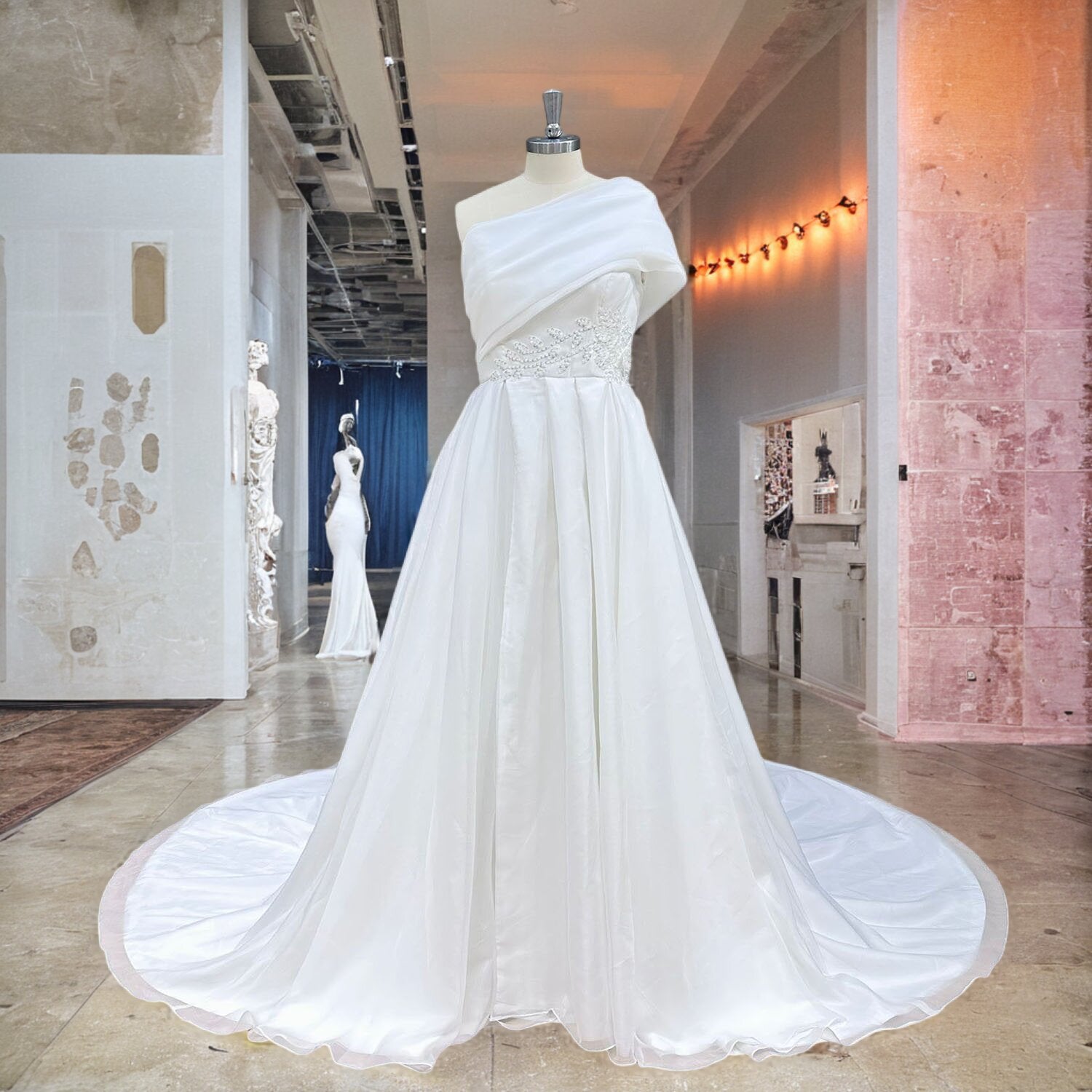 Front view of a white asymmetric one-shoulder bridal gown on a mannequin, showcasing a softly draped bodice and intricate silver beadwork cascading along the wearer's right hip.