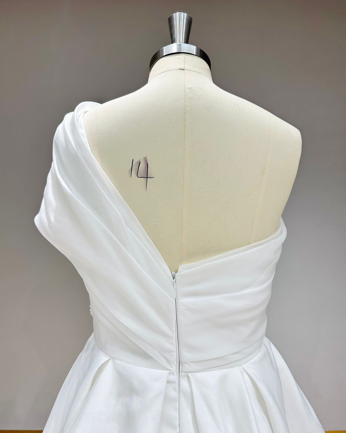 Detailed upper back view of a minimalist one-shoulder wedding gown on a dress form, highlighting the clean fabric wrap and a hidden side zipper closure.
