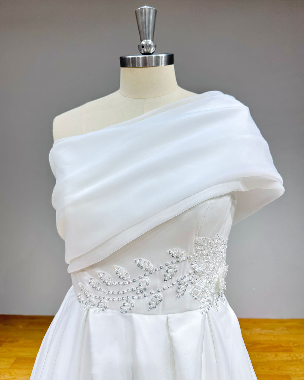Close-up detail of a white bridal bodice showcasing an asymmetric sheer organza shoulder overlay and a waistband encrusted with leaf motifs and silver sequins.