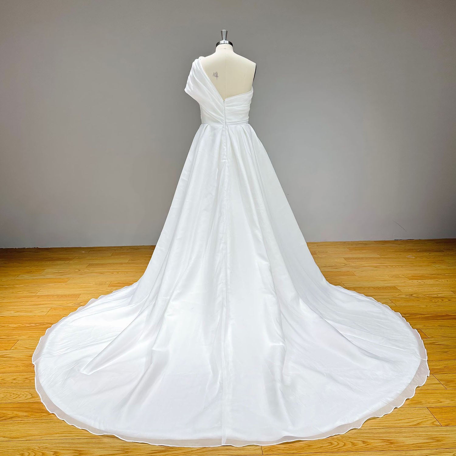 Studio back view of a Grecian style one shoulder wedding gown on a mannequin, showcasing a sweeping chapel-length organza train that drapes smoothly to the floor.