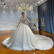 Full front view of a model wearing a sparkling luxury beaded bridal dress featuring sheer long sleeves, an illusion neckline, and a dramatic, voluminous ball gown skirt with a long train.