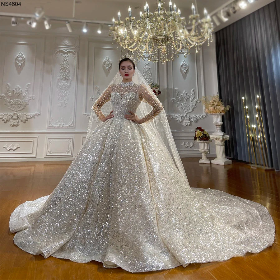 Full-length front view of a bride wearing a sparkling luxury princess ball gown wedding dress with long illusion sleeves, heavy crystal beadwork, and a voluminous cathedral train.