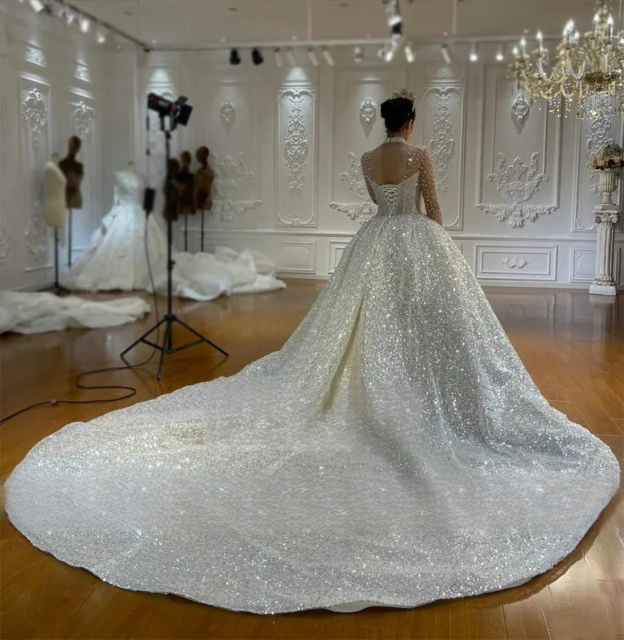Dramatic rear view of a bride in a sparkling ball gown wedding dress, showcasing the classic lace-up corset back and the expansive, shimmering silver sequin train spreading across the floor.
