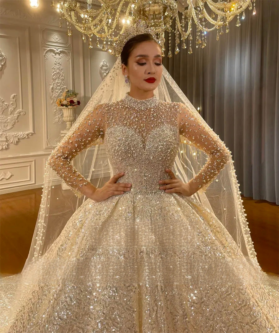 Front-facing close-up of a bride in a luxury ball gown wedding dress, emphasizing the high jewel neckline, intricate silver beadwork on sheer sleeves, and a matching pearl-trimmed veil.