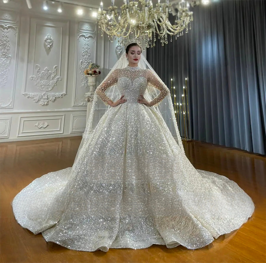 Full-length front view of a bride posing in a long-sleeve sparkling ball gown wedding dress, showcasing the uniform crystal embellishment and regal, structured silhouette.