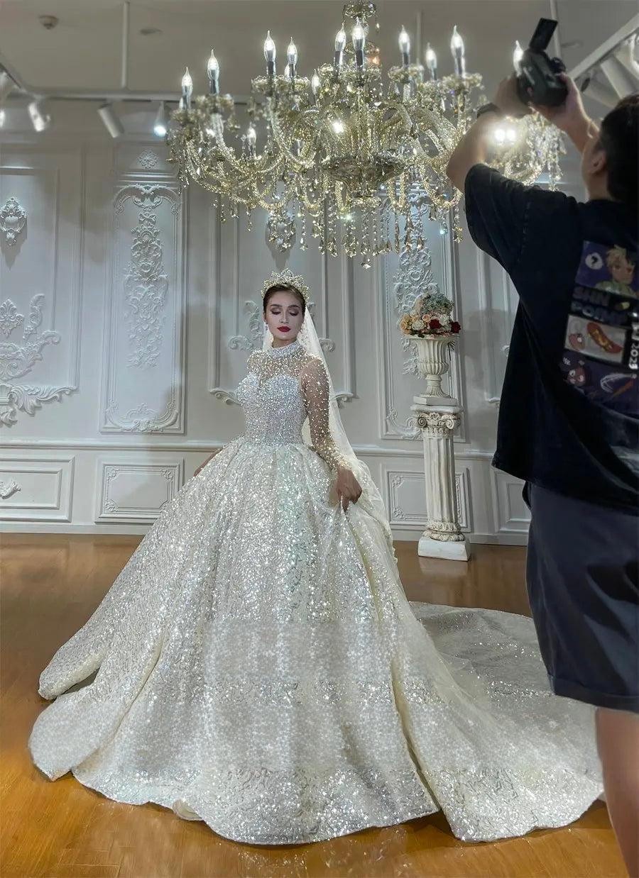Behind-the-scenes shot of a bride modeling a luxury sparkling ball gown wedding dress during a photoshoot under an ornate crystal chandelier.