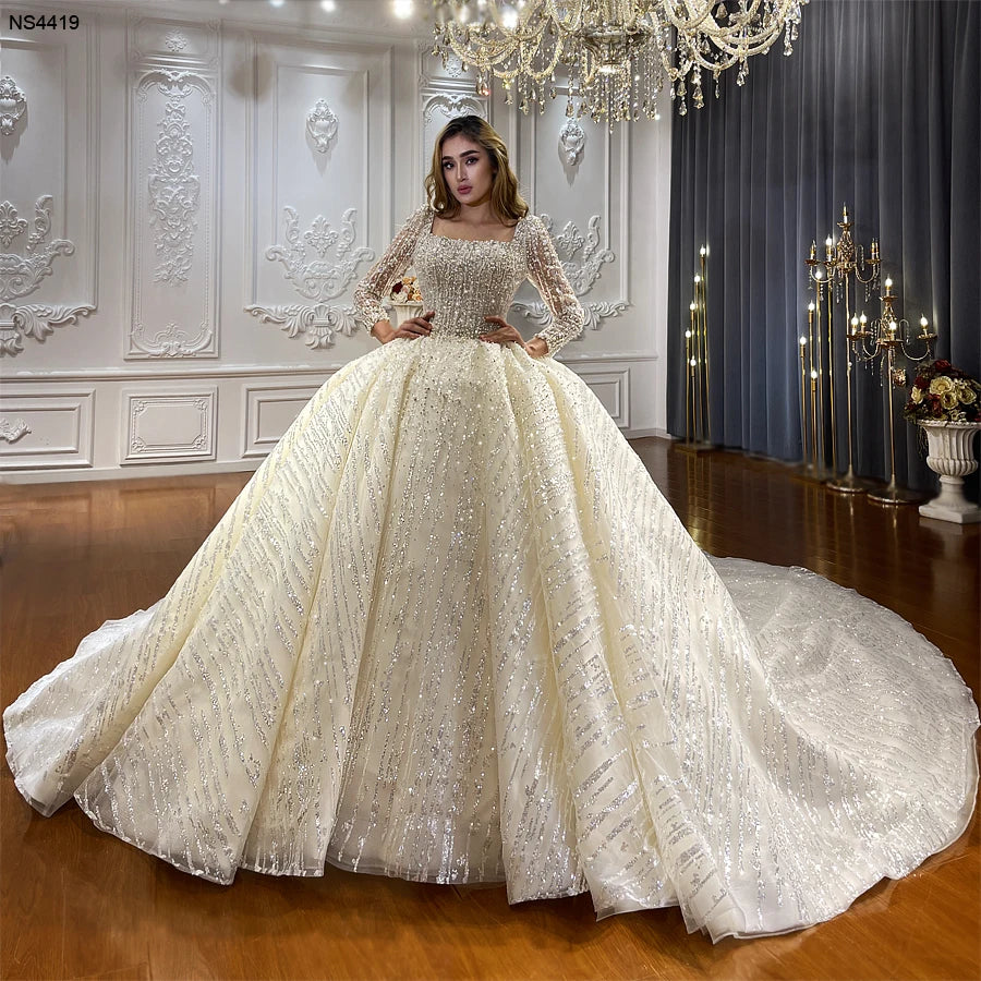 Full front view of a luxury square neck long sleeve ball gown wedding dress featuring shimmering vertical crystal patterns and a grand fluffy voluminous skirt.