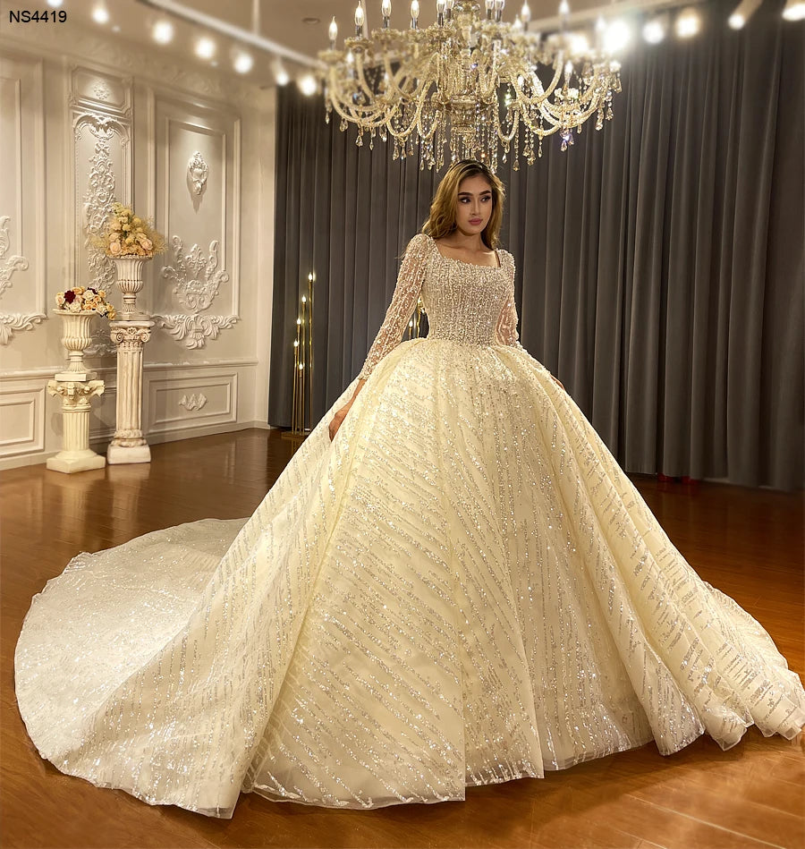 Elegant front pose of a sparkling square neck bridal gown with sheer long sleeves and a royal-style fluffy ball gown silhouette in a professional studio.