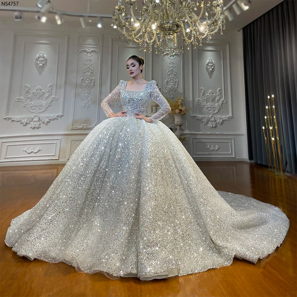 Full-length front view of a luxury square neckline long sleeve sparkly ball gown wedding dress with an expansive sequined skirt and shimmering silver details in a grand bridal showroom.