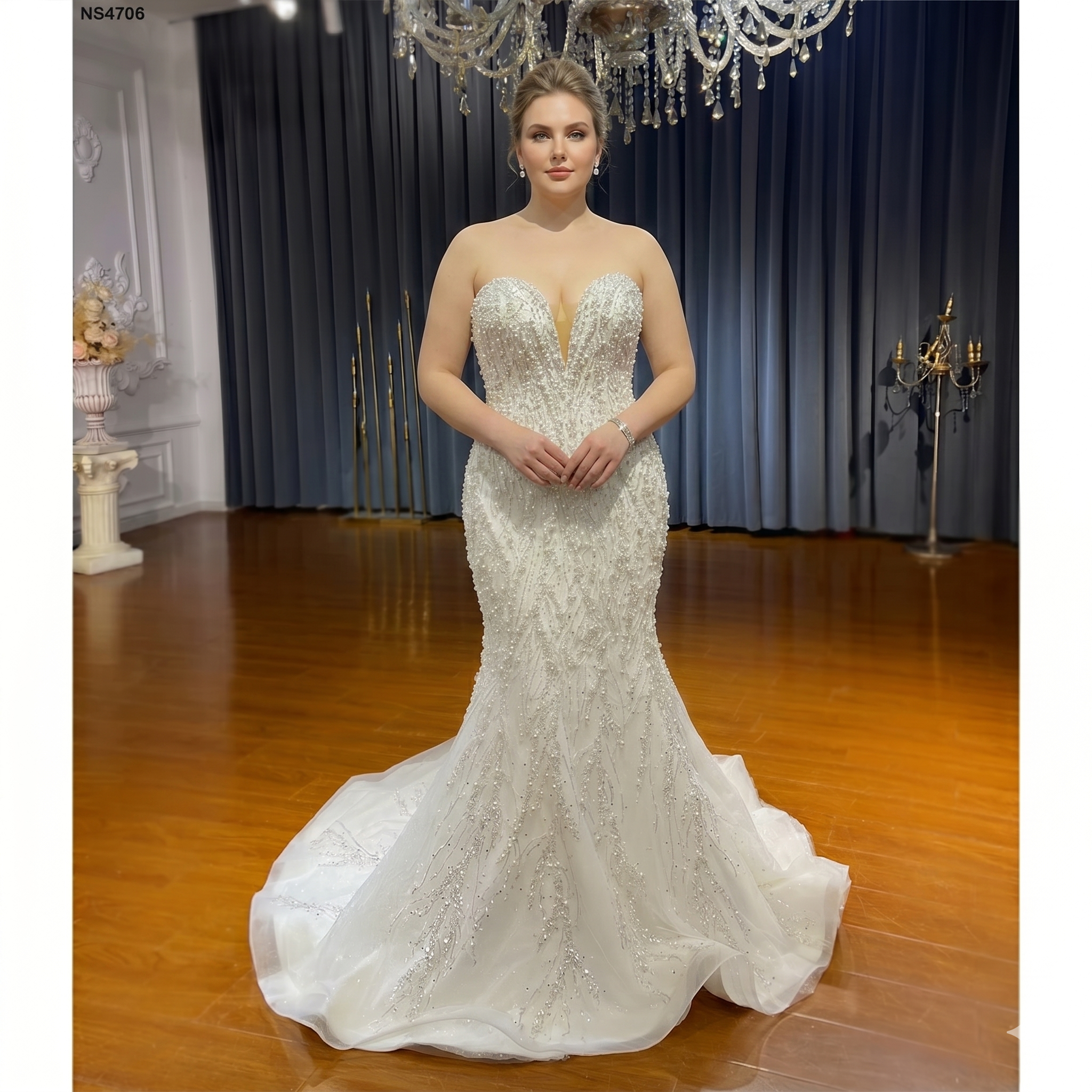 A plus-size model wearing an elegant strapless mermaid bridal gown featuring heavy pearl embellishments and a plunging sweetheart neckline with illusion mesh.