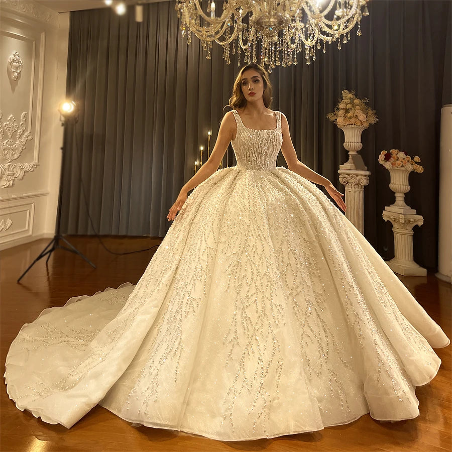 Angled front perspective of an elegant model presenting a luxury thick strap ball gown wedding dress, highlighting the regal silhouette and dense, shimmering crystal embroidery under chandelier lighting.