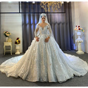 Full front view of a bride wearing a stunning off-white bridal gown featuring heavy beading, a sheer illusion neckline, long sleeves with 3D floral appliqués, and a voluminous ball gown skirt.