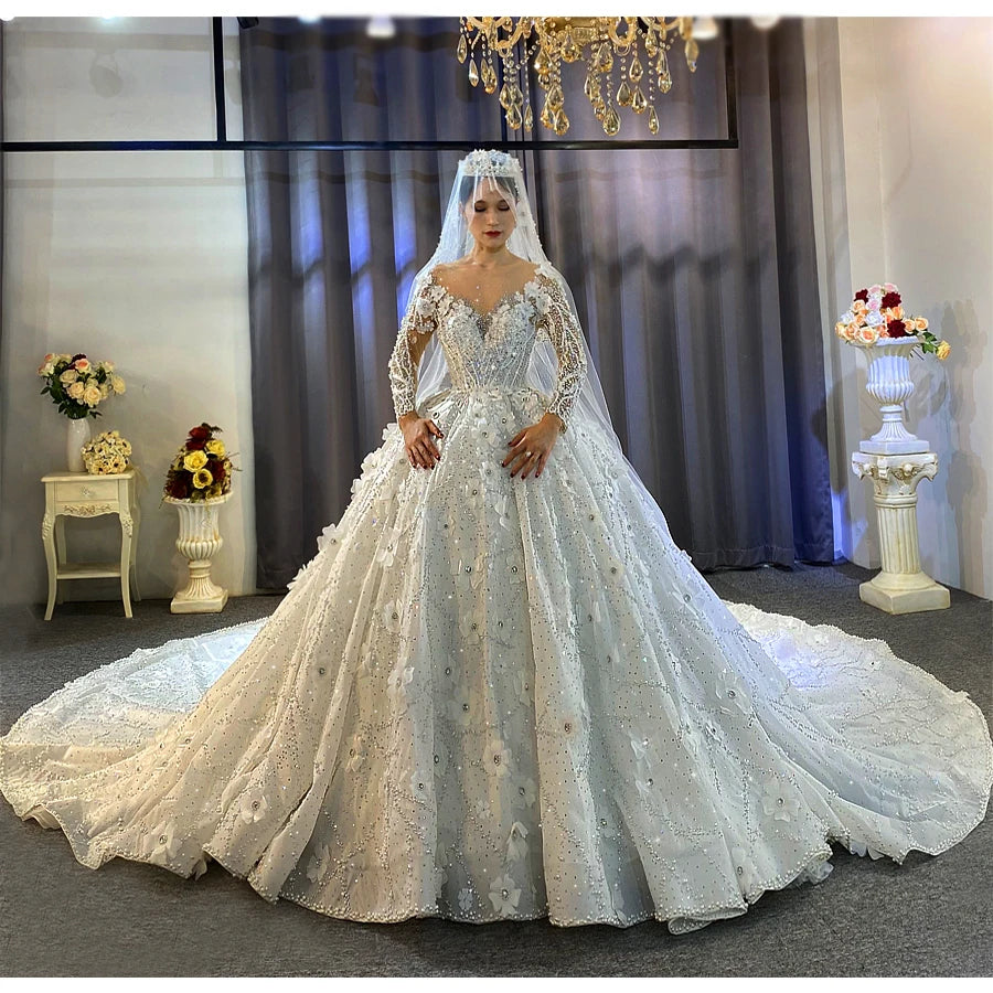 Full front view of a bride wearing a stunning off-white bridal gown featuring heavy beading, a sheer illusion neckline, long sleeves with 3D floral appliqués, and a voluminous ball gown skirt.