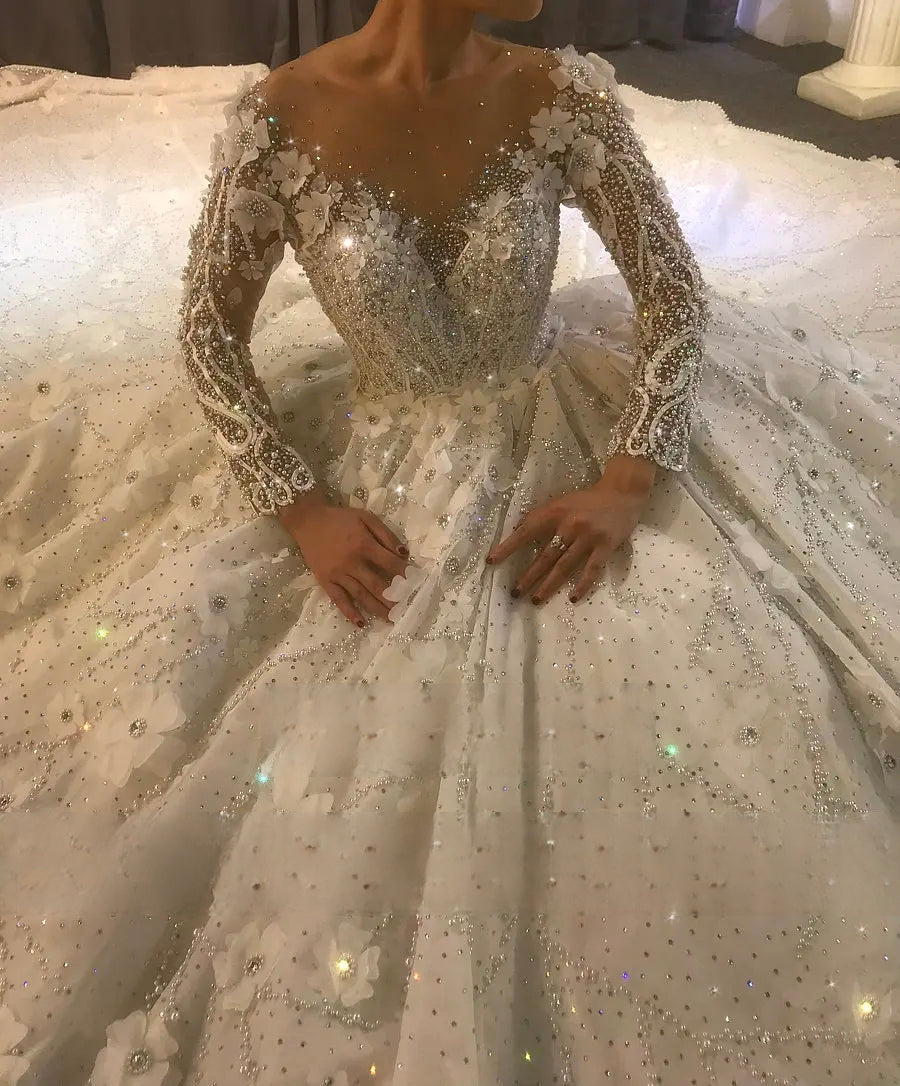 High-angle close-up of the elegant wedding dress bodice, emphasizing the sheer illusion neckline, heavy crystal beading, and intricate 3D floral lace on the long sleeves.