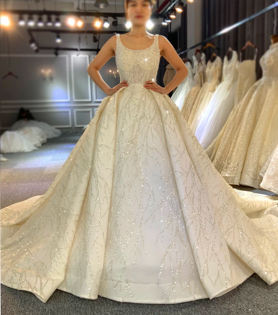 Full-length front view of a bride modeling a stunning sleeveless luxury ball gown with full beaded embellishments and a deep scoop neckline in a brightly lit bridal studio.