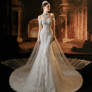 Full-length front view of an elegant model wearing a stylish mermaid wedding dress featuring off-the-shoulder cap sleeves with beaded tassels, heavy crystal embellishments, and a cascading sheer tulle cape train.