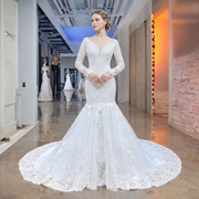 Elegant model wearing a long-sleeved sweetheart mermaid wedding dress with intricate pearl lace and a sweeping circular train in a luxury bridal showroom.