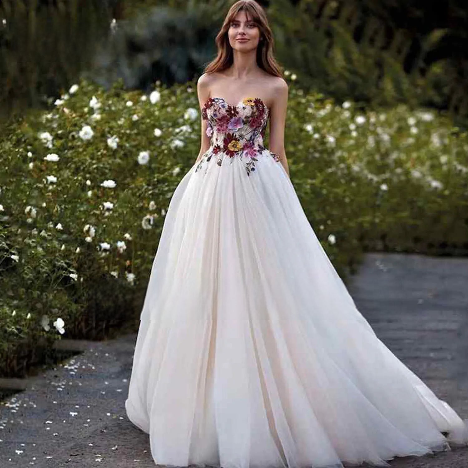 An elegant bride modeling a colorful floral wedding dress outdoors, standing in a garden setting wearing a sweetheart neck ball gown with vibrant 3D flower appliques.
