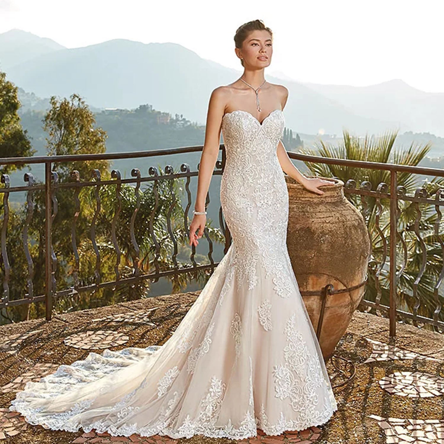 Bride posing on a scenic mountain overlook terrace wearing a strapless lace mermaid wedding gown with a soft champagne underlay and flowing train.