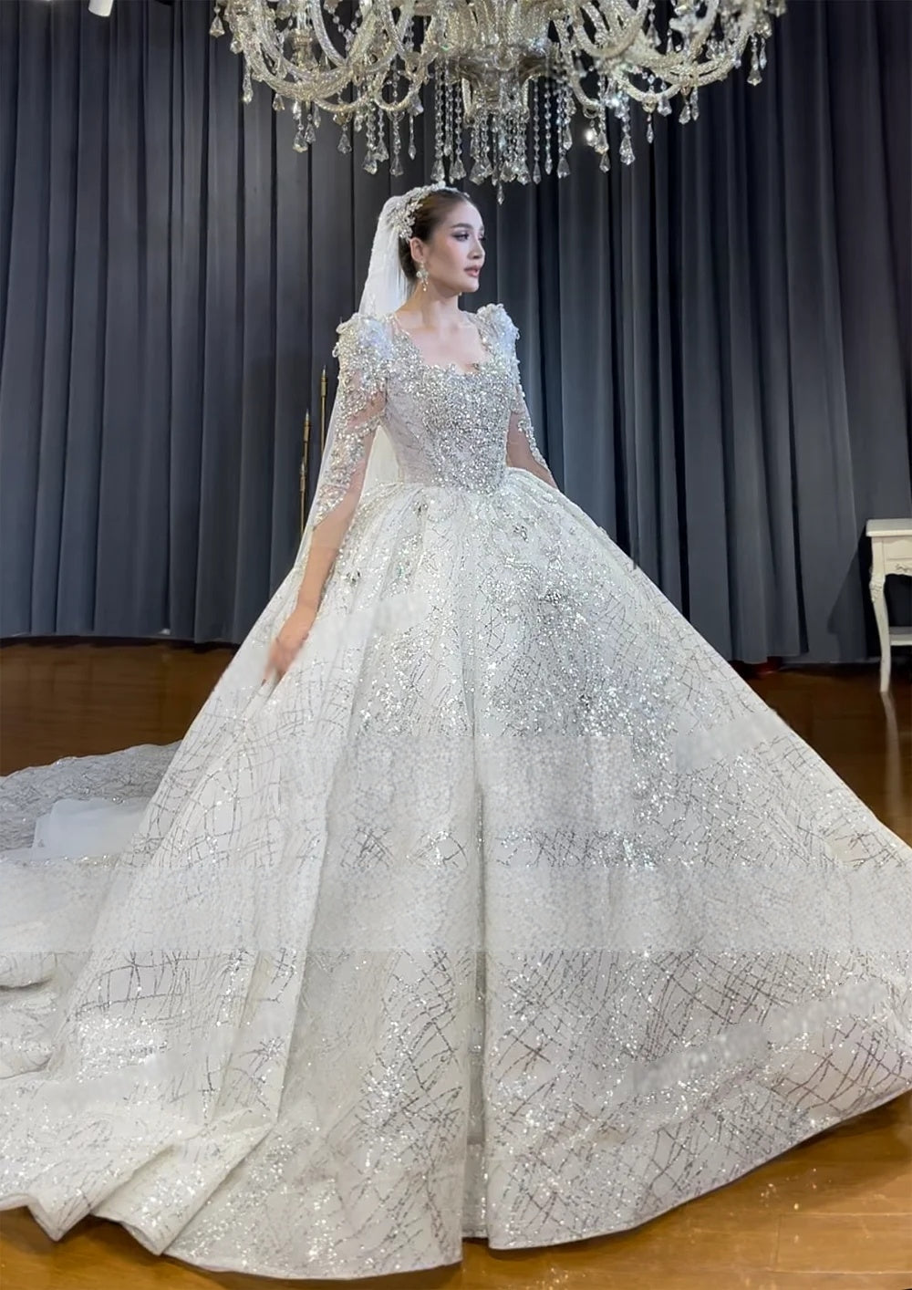 Front-side angle of a voluminous beaded ball gown wedding dress with long sleeves, showcasing the shimmering fabric and grand presence in a luxury bridal studio.
