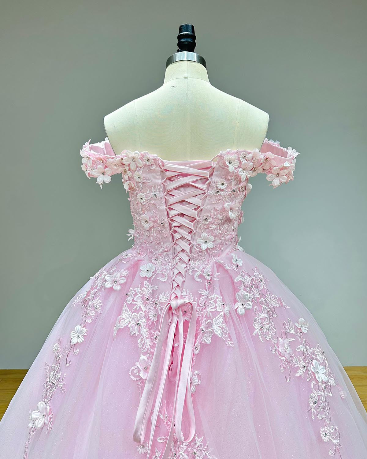Close-up back bodice view of a sweetheart pink dress on a mannequin, showcasing the traditional lace-up back detail framed by elegant 3D flowers, pearls, and crystals along the off-shoulder straps.