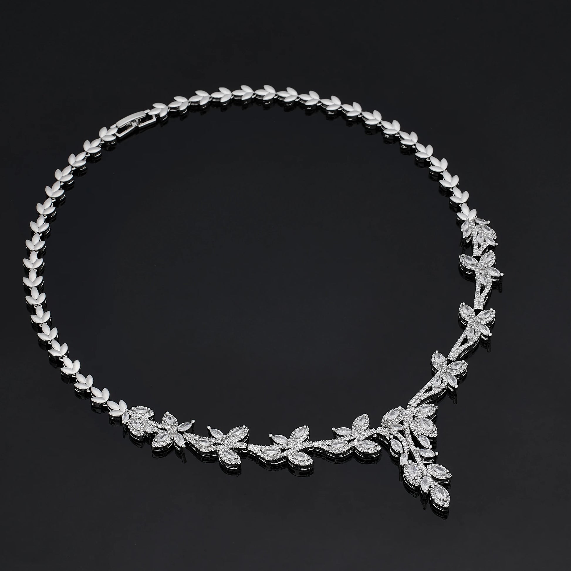 Overhead view of a silver branch-motif cubic zirconia wedding necklace, highlighting its detailed leafy pave structure on a dark reflective surface.
