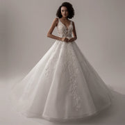 A model wearing an ivory, sleeveless, V-neck A-line wedding dress with intricate lace appliques, standing in a studio.