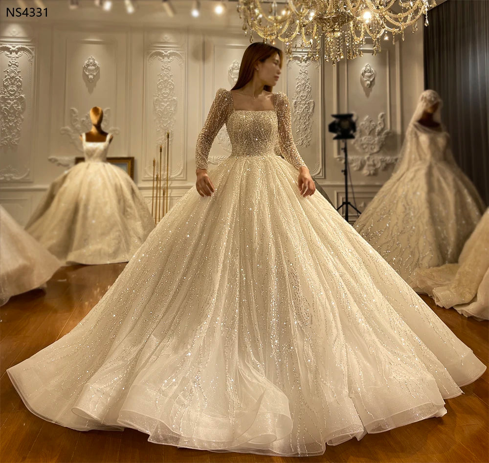 Full-length side view of a model wearing an elegant vintage long sleeve beaded ball gown wedding dress, featuring a massive sparkling skirt with cascading sequin patterns in a luxury showroom.