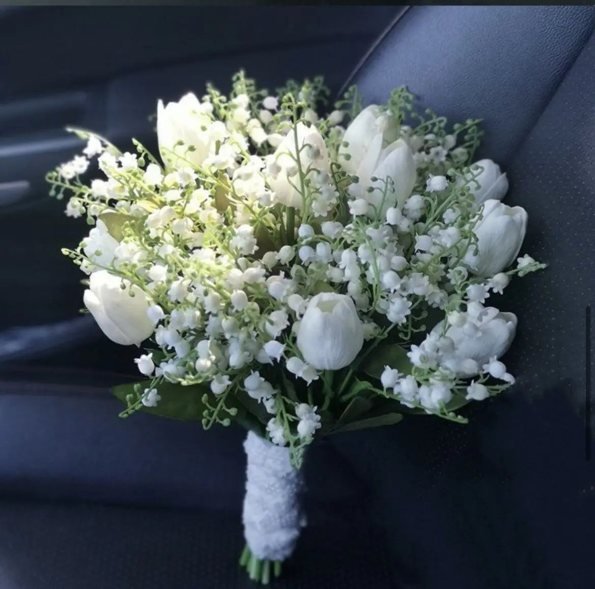 Close-up of a handmade artificial bridal bouquet featuring white tulips and lily of the valley with a lace-wrapped handle.