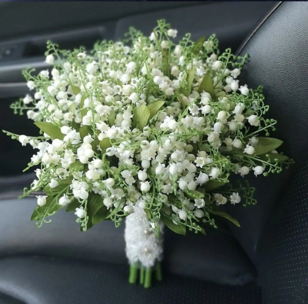 Detailed top view of a handmade artificial wedding bouquet made of white lily of the valley and greenery with a lace-wrapped handle.