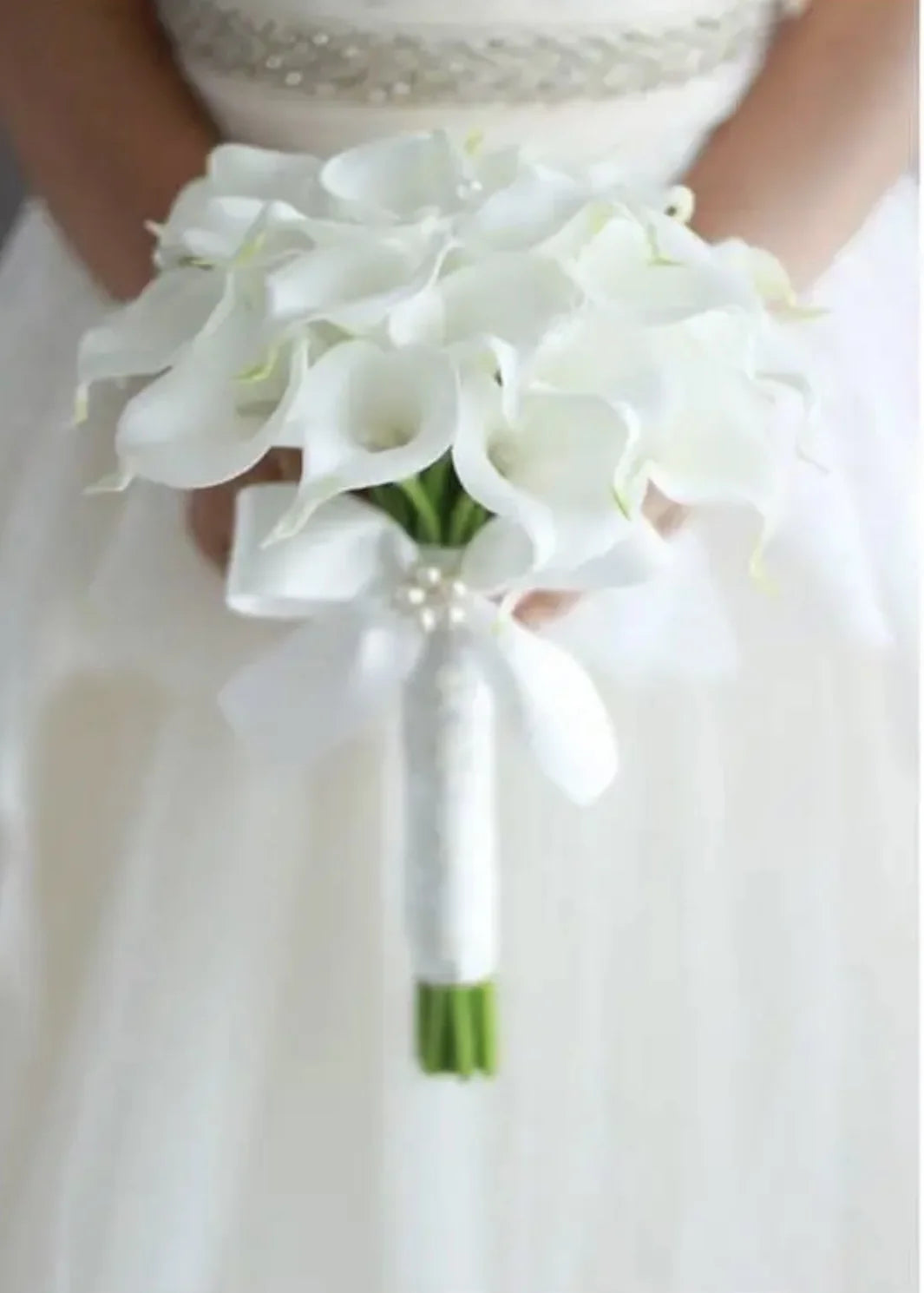 A bride holding a delicate, handmade white calla lily wedding bouquet accented with a white ribbon and pearls.