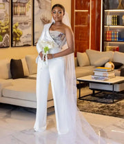 Full-length front view of a bride wearing a modern white one-shoulder wedding jumpsuit featuring a strapless metallic embellished bodice, a structured asymmetric shoulder drape, tailored wide-leg trousers, and a long trailing tulle veil.