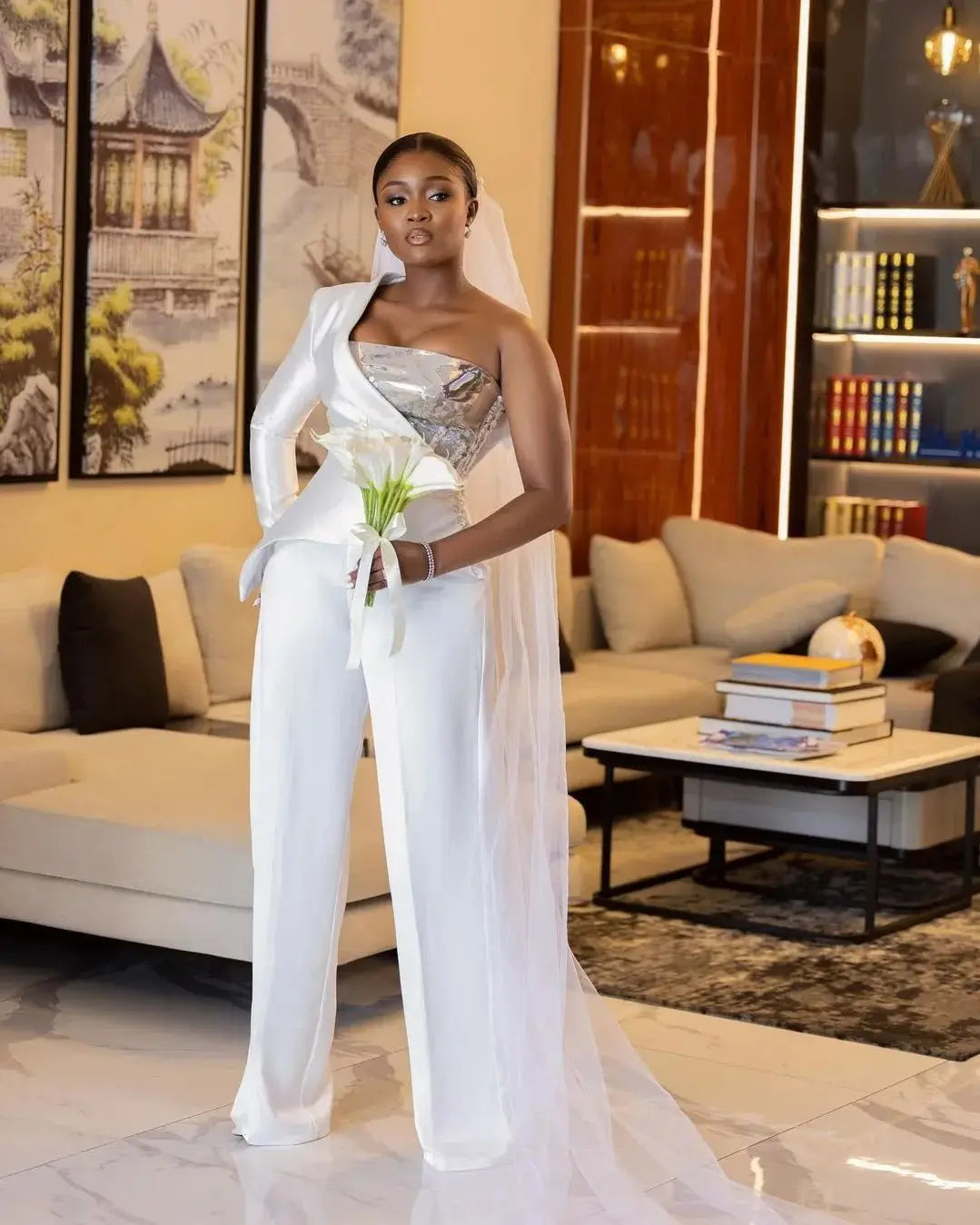 Front shot of a bride in a plus size one-shoulder strapless bridal pantsuit, highlighting the unique asymmetric satin drape, shimmering silver corset top, and a white calla lily bouquet.
