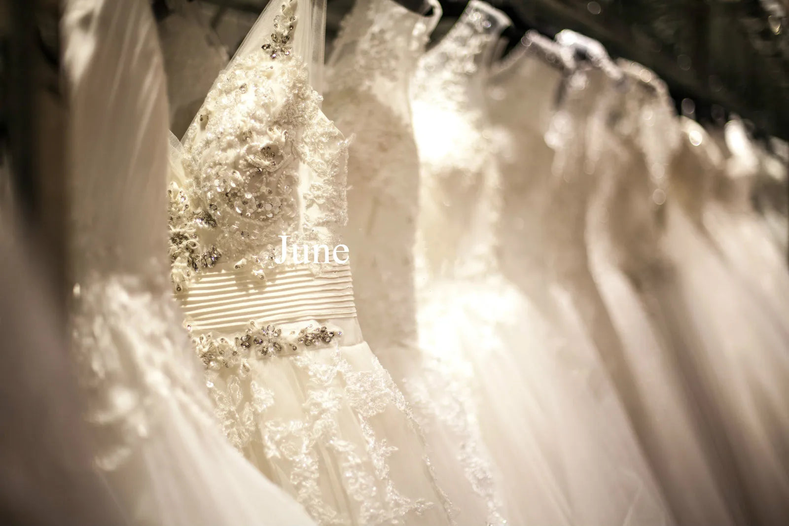 A close-up, softly focused view of heavily beaded and embroidered white wedding dresses hanging on a rack in a bridal boutique with a text overlay that reads "June".
