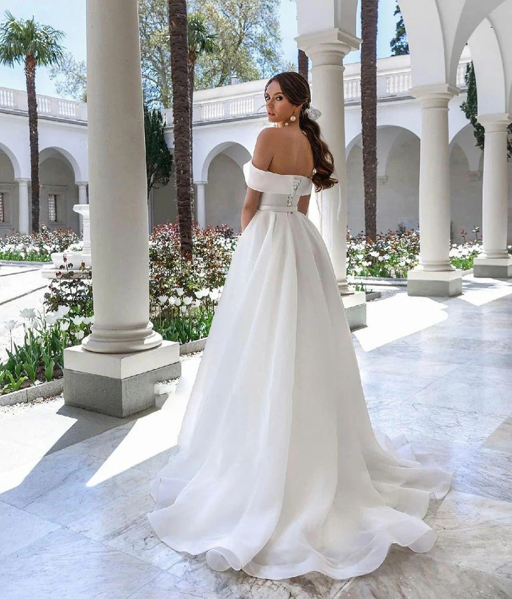 Back view of a white organza A-line bridal gown with a sophisticated off-the-shoulder style in a scenic garden setting.