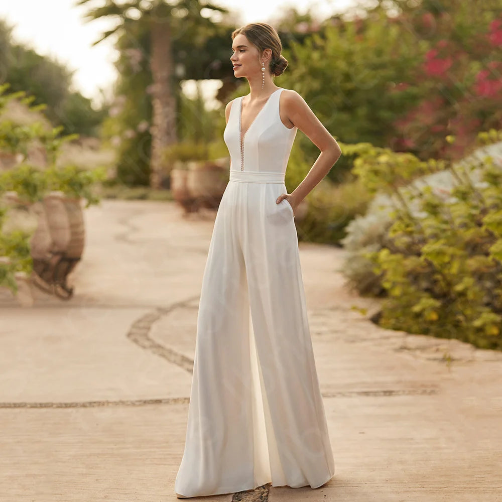 Full-length front view of a bride posing on an outdoor pathway in a sleeveless white wedding jumpsuit featuring a plunge V-neckline with delicate center trim and relaxed wide-leg trousers with pockets.